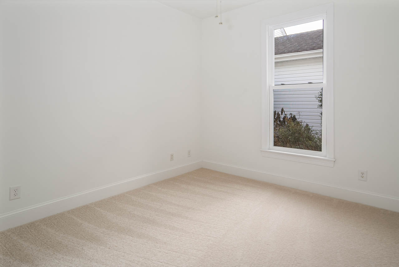 A small, empty room with beige carpet, white walls, and a single window letting in natural light. The window shows part of a white building and some plants outside. There are several electrical outlets along the walls.