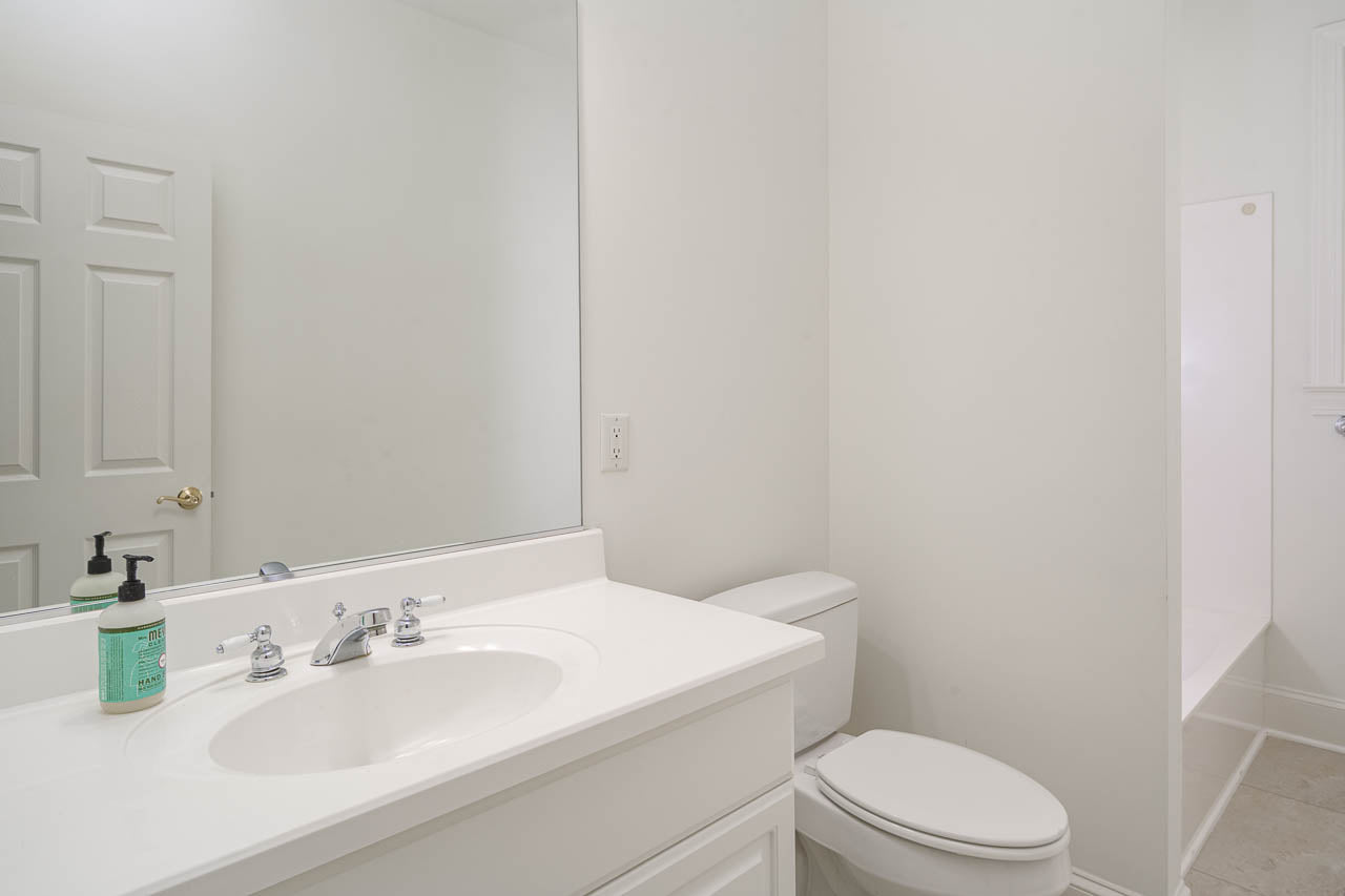 A clean, white bathroom with a sink, faucet, soap dispensers, toilet, large mirror, and a walk-in shower visible in the background. The walls and fixtures are all white, creating a bright, minimalist look.