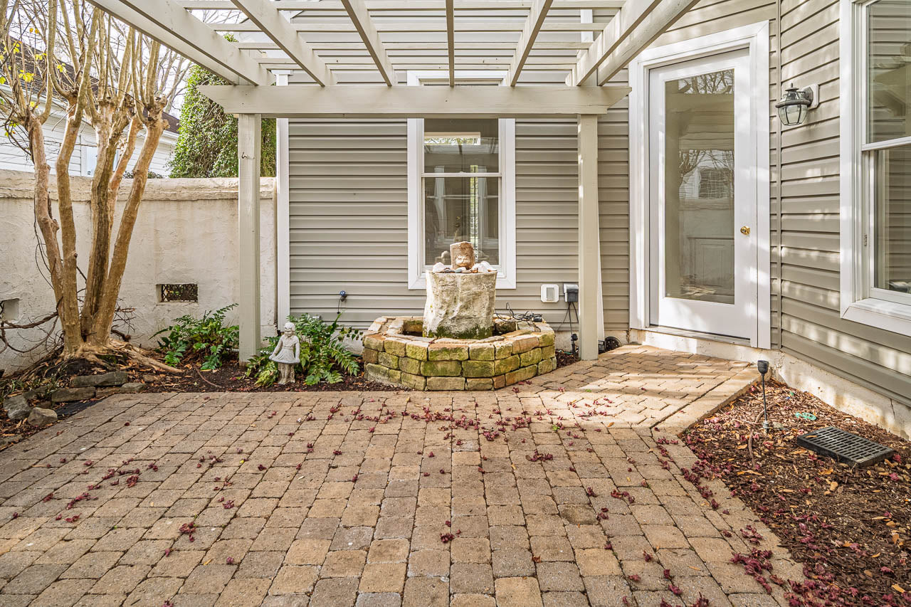 A small, paved patio features a central stone fountain, surrounded by a low brick border and plants, with a pergola overhead and a door leading into a gray-sided house. Fallen leaves are scattered on the paving stones.