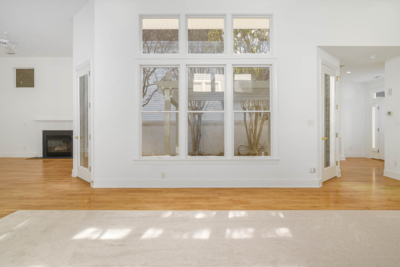 Bright, empty living room with large windows, hardwood floors, a cream-colored rug, glass doors, and a view of an outdoor pergola and wall through the windows. White walls and high ceilings create an airy feel.