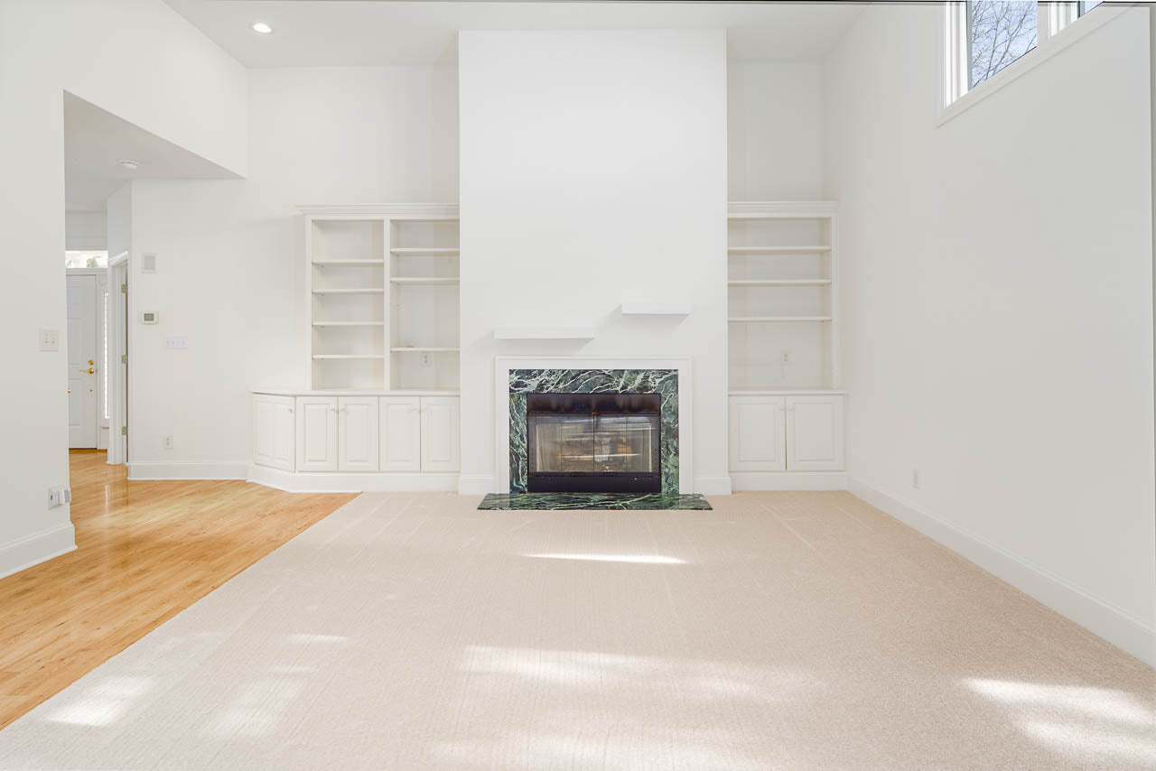 Bright, empty living room with white walls, built-in white shelves on either side of a fireplace with a dark green marble surround, light carpet, and hardwood flooring. Large windows let in natural light.