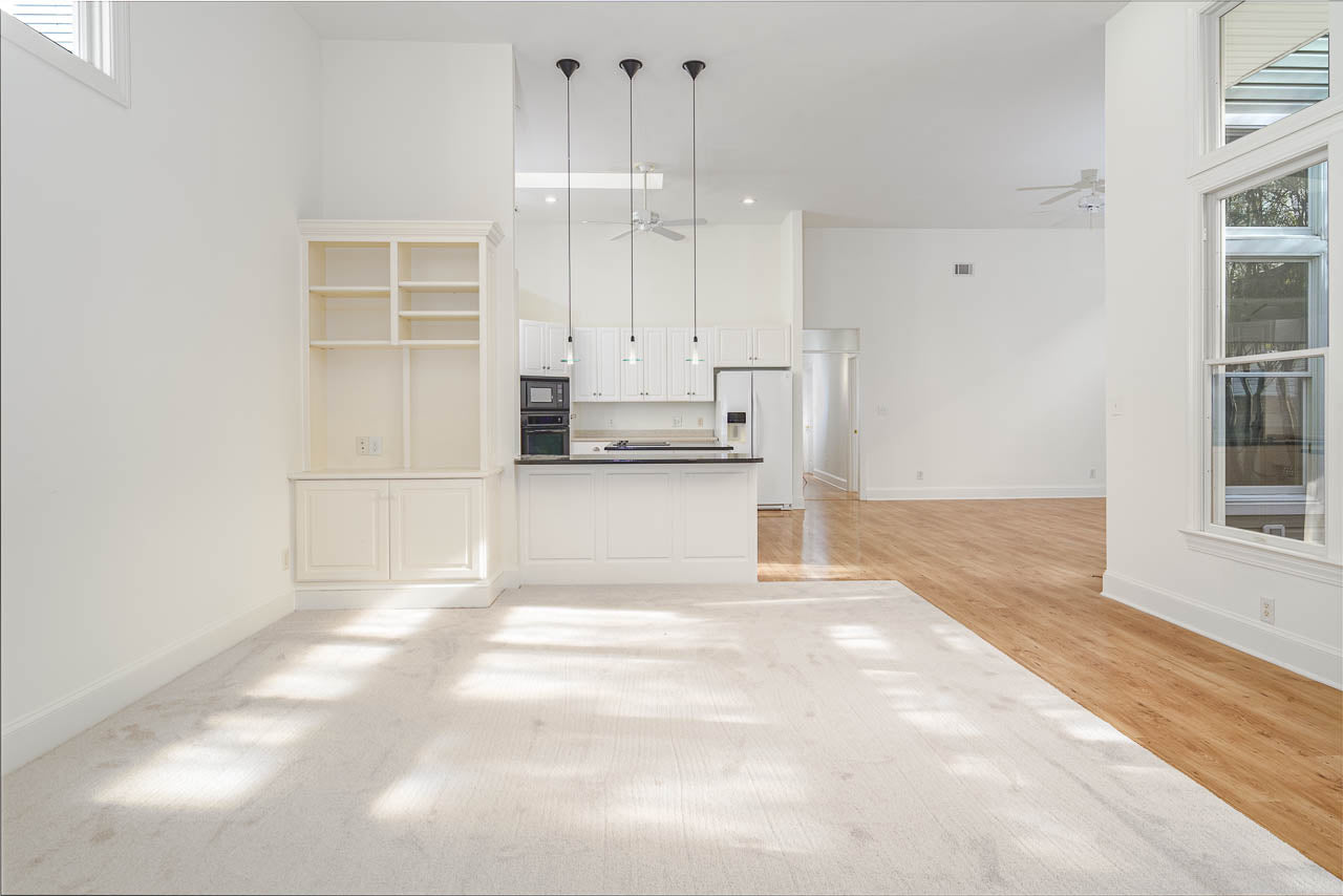 A bright, spacious open-plan room with white walls and cabinetry, a kitchen island with pendant lights, hardwood flooring, and large windows letting in natural light. The carpeted area meets the hardwood floor near the kitchen.
