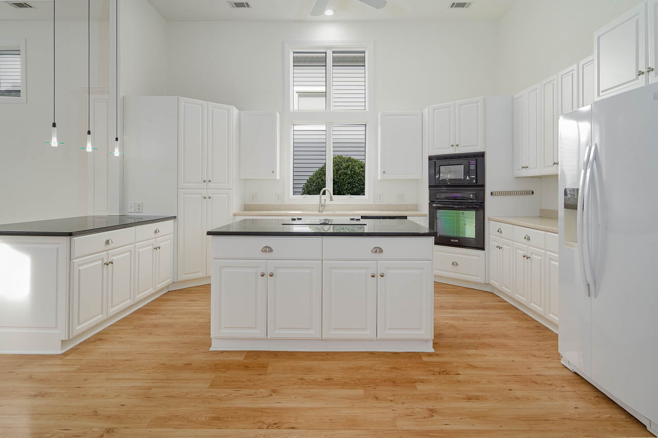 Spacious modern kitchen with white cabinets, black countertops, wood flooring, built-in double ovens, large refrigerator, island with sink, and tall windows letting in natural light.