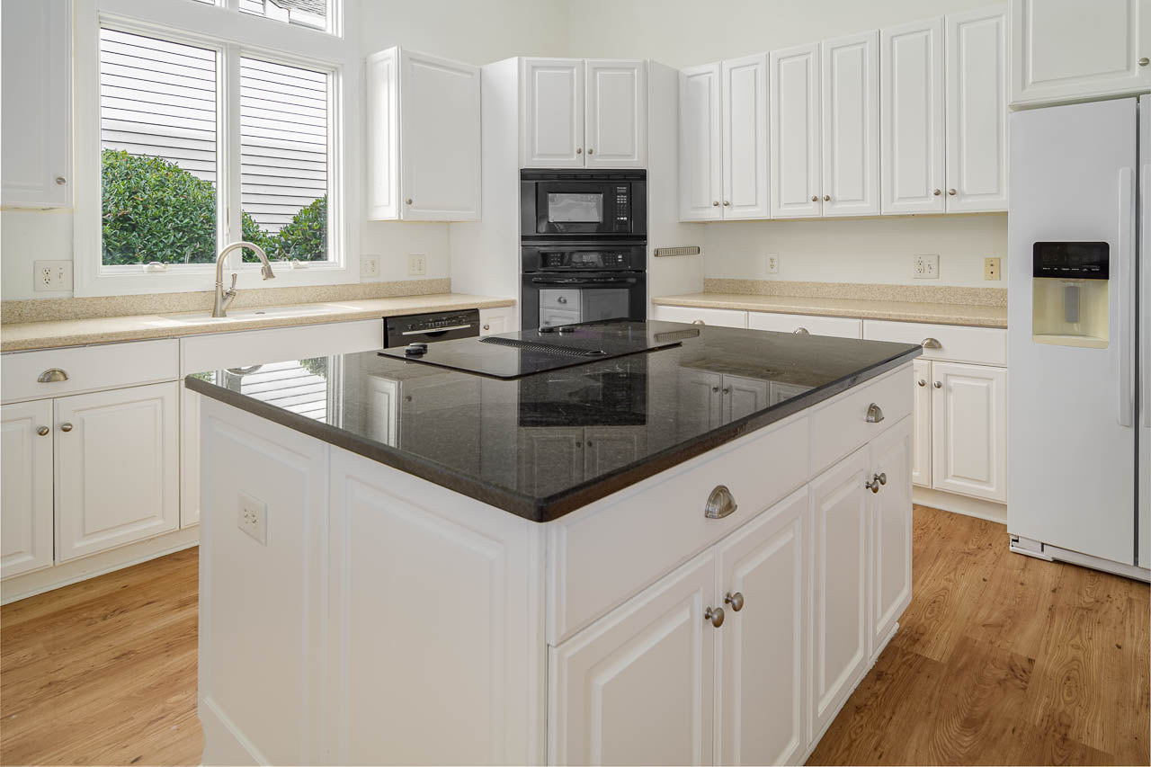 A modern kitchen with white cabinets, a large black granite island, built-in black appliances, a sink under a window, a white refrigerator, and light wood flooring.