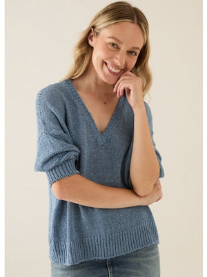 A woman with blonde hair models the BASE MILANO - V NECK COTTON SWEATER in light blue, paired with matching jeans, standing against a plain light background and gazing slightly to the side.