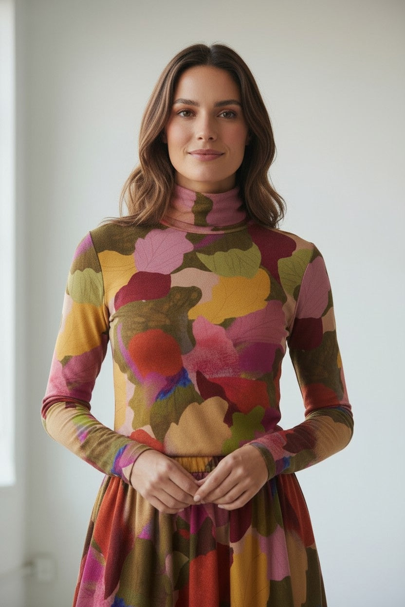 A woman stands indoors wearing the FRANCO FERRARI FRAGOLINA TURTLENECK, a long-sleeve dress featuring a bold abstract floral and leaf pattern in vibrant red, pink, yellow, and green. She has wavy brown hair and smiles softly at the camera.