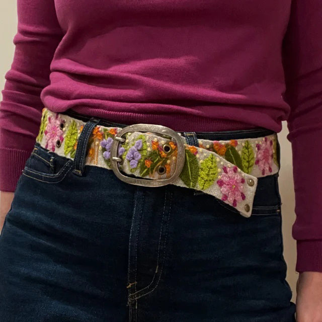 A person wears a magenta long-sleeve top, dark blue jeans, and the JENNY KRAUSS - FRESHWATER BELT by JENNY KRAUSS, handcrafted in Peru and adorned with colorful embroidered flowers and leaves.