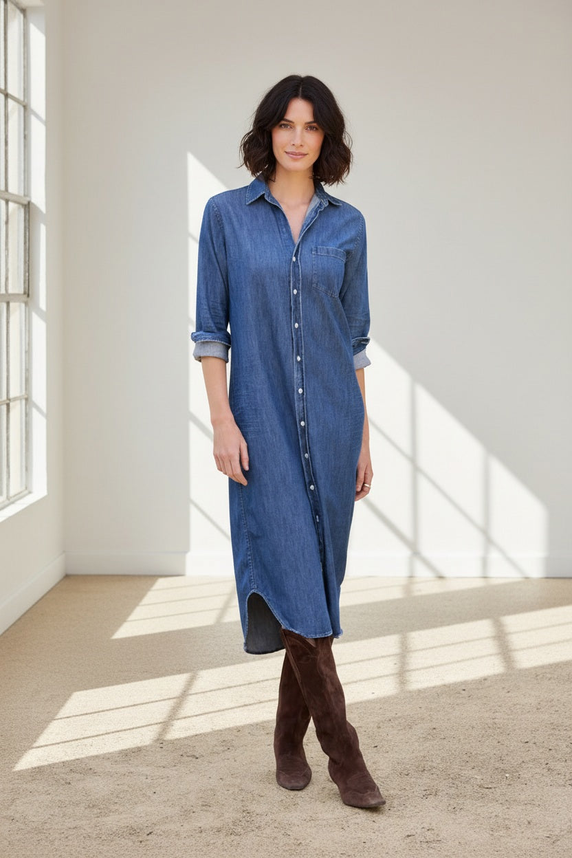 A woman stands outdoors in the FRANK & EILEEN Rory Maxi Shirtdress in Distressed Indigo Washed Denim, its long button-up silhouette and rolled sleeves crafted from luxurious indigo fabric. She pairs the effortlessly chic dress with brown knee-high boots, set against a backdrop of lush greenery and a building.