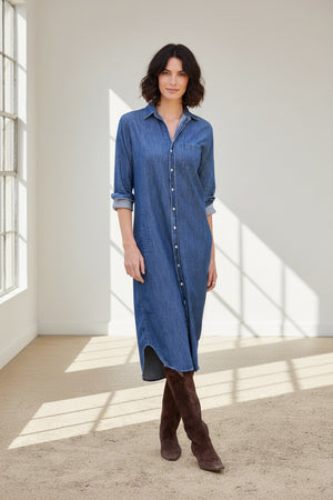 A woman stands outdoors in the FRANK & EILEEN Rory Maxi Shirtdress in Distressed Indigo Washed Denim, its long button-up silhouette and rolled sleeves crafted from luxurious indigo fabric. She pairs the effortlessly chic dress with brown knee-high boots, set against a backdrop of lush greenery and a building.