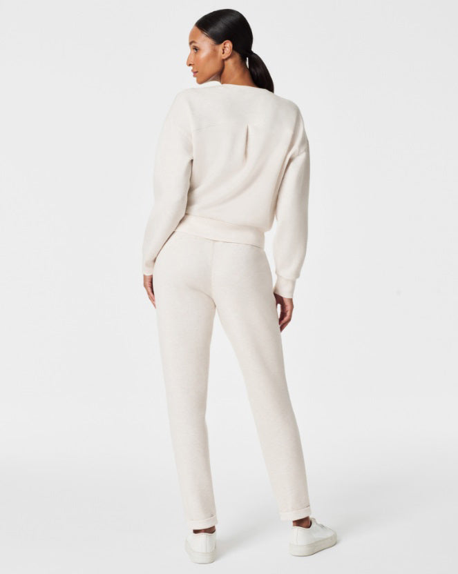 A person stands against a white background, smiling and looking down with one hand touching their hair. They are wearing a matching light beige SPANX AIRESSENTIALS TAPERED PANT and sweatshirt set made from ultra drapey, lightweight spacer fabric, paired with white sneakers.