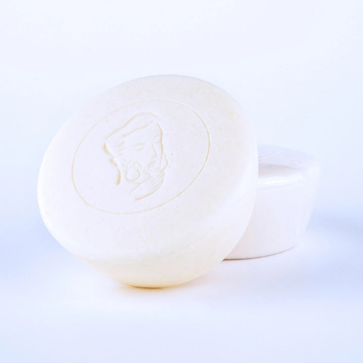 Two round, white BAUDELAIRE - M.B. SHAVING SOAP bars by BAUDELAIRE are displayed. The front bar features an embossed profile of a bearded man, set against a plain white background that evokes the clean, minimalist feel and hints at the smooth razor glide and nourishing shea butter found in each bar.