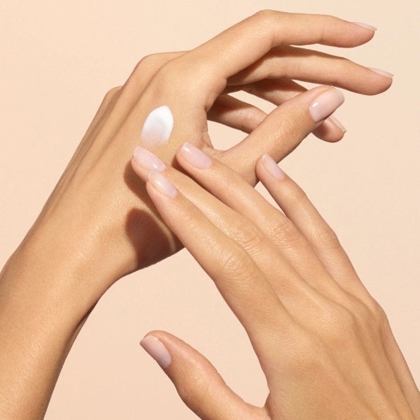 Close-up of two hands with light skin; one is gently applying MANUCURIST - COSMOS ORGANIC HAND CREAM, ORANGE BLOSSOM by MANUCURIST INC to the back of the other against a neutral beige background. Ideal for hand and nail care, this organic moisturizer is formulated with natural ingredients to leave fingers nourished. The nails are perfectly groomed and finished with a subtle pale nude polish for an elegant touch.