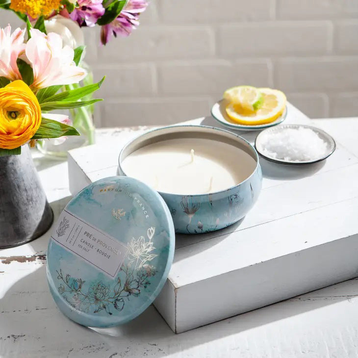 The 3 Wick Candle - Sea Salt by European Soaps, LLC comes in a round tin adorned with a pale blue watercolor design featuring floral and botanical illustrations. The text reads &quot;Parfum de France, Pré de Provence, Candle Bougie, Sea Salt, 10.5 oz/300g,&quot; and the design includes an illustration of a bird at the top. This candle is ideal for enhancing home ambience with its sparkling aldehydes scent.