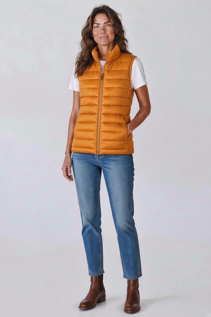 A woman wearing a SAVE THE DUCK - LYNN PUFFER VEST IN AMBER ORANGE over a white t-shirt, blue jeans, and brown ankle boots.