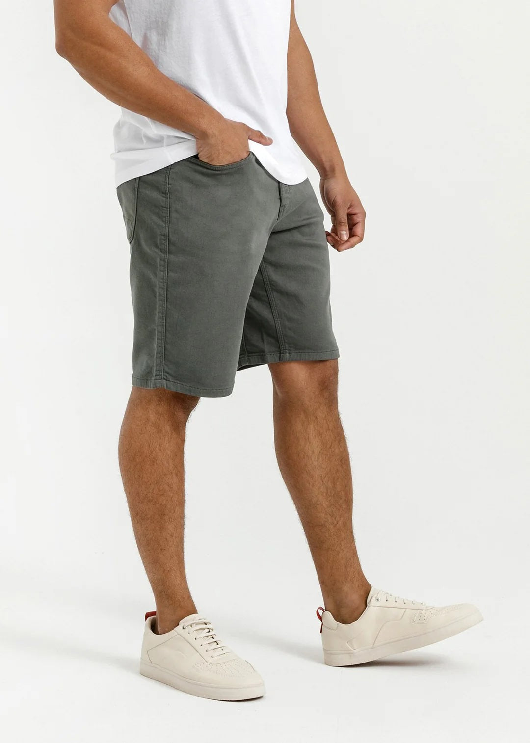 A person stands against a plain light background, wearing a white T-shirt, DUER No Sweat Relaxed Shorts in Gull made from stretch fabric, and white sneakers, with one hand in their pocket.