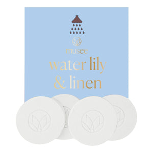 A blue box labeled “MUSEE - WATER LILY & LINEN SHOWER STEAMERS” from MUSEE sits behind five round white tablets, each embossed with a delicate leaf design. These shower steamers offer a spa-like escape, infused with the gentle scent of water lily. Above the text on the box, a small showerhead icon is featured.