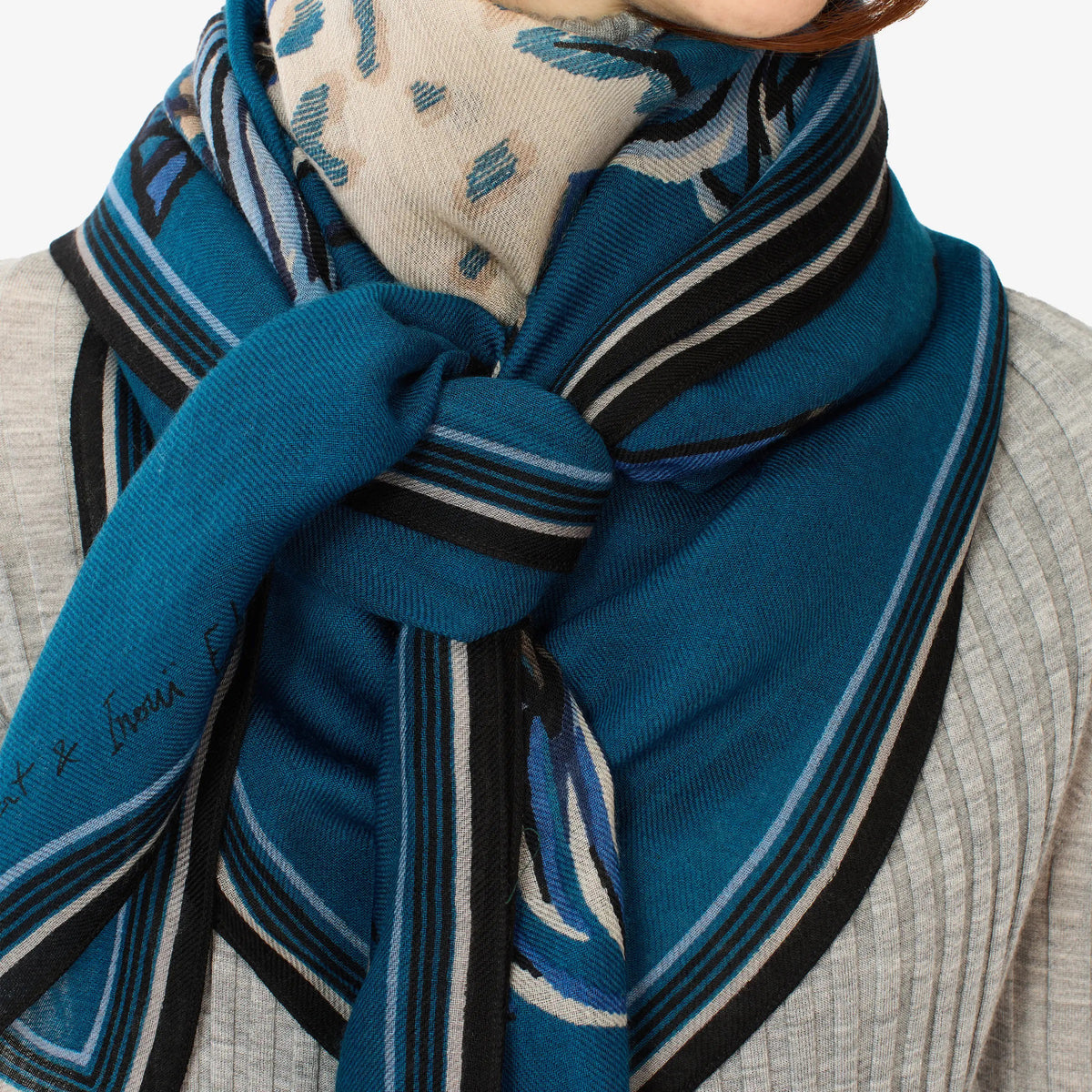 A person with straight, reddish-brown hair models the INOUI EDITIONS - WESTERN SQUARE SCARF by INOUI, a blue and white patterned wool accessory made in India, draped over a light gray ribbed sweater and black pants, standing against a plain white background.