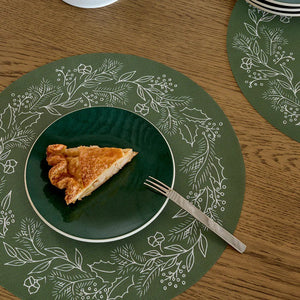 A slice of pie on a dark green plate with a fork rests on the CHILEWICH - WREATH PRINT PLACEMAT by CHILEWICH, featuring white botanical designs that add charm to the wooden table.