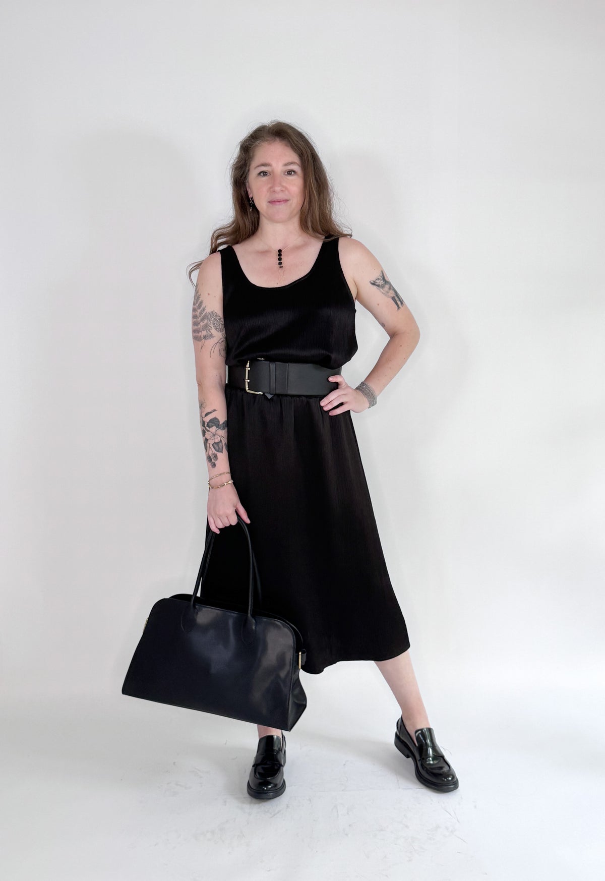 A woman in a sleeveless black dress with a wide belt stands against a plain backdrop, holding a large black handbag. She wears black shoes and layers the ROSE AND CROWN - SATIN RIBBED SKIRT by ROSE &amp; CROWN under her dress.
