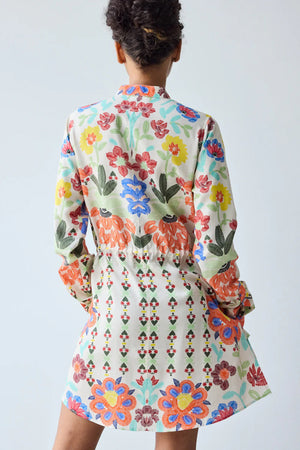 A woman with short curly hair and tattoos on her left leg stands indoors by a plain wall, wearing the HOPE FOR FLOWERS - BAND COLLAR TUNIC IN CUSTARD FLORAL from HOPE FOR FLOWERS, paired with sandals.