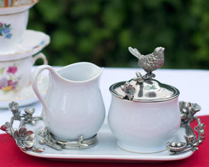 The VAGABOND HOUSE - SONG BIRD CREAMER SET by VAGABOND HOUSE includes a white ceramic sugar bowl and creamer, each with ornate silver floral handles and a bird-shaped lid handle. The elegant set also features a decorative tray and a small spoon with a floral handle for stylish serving.