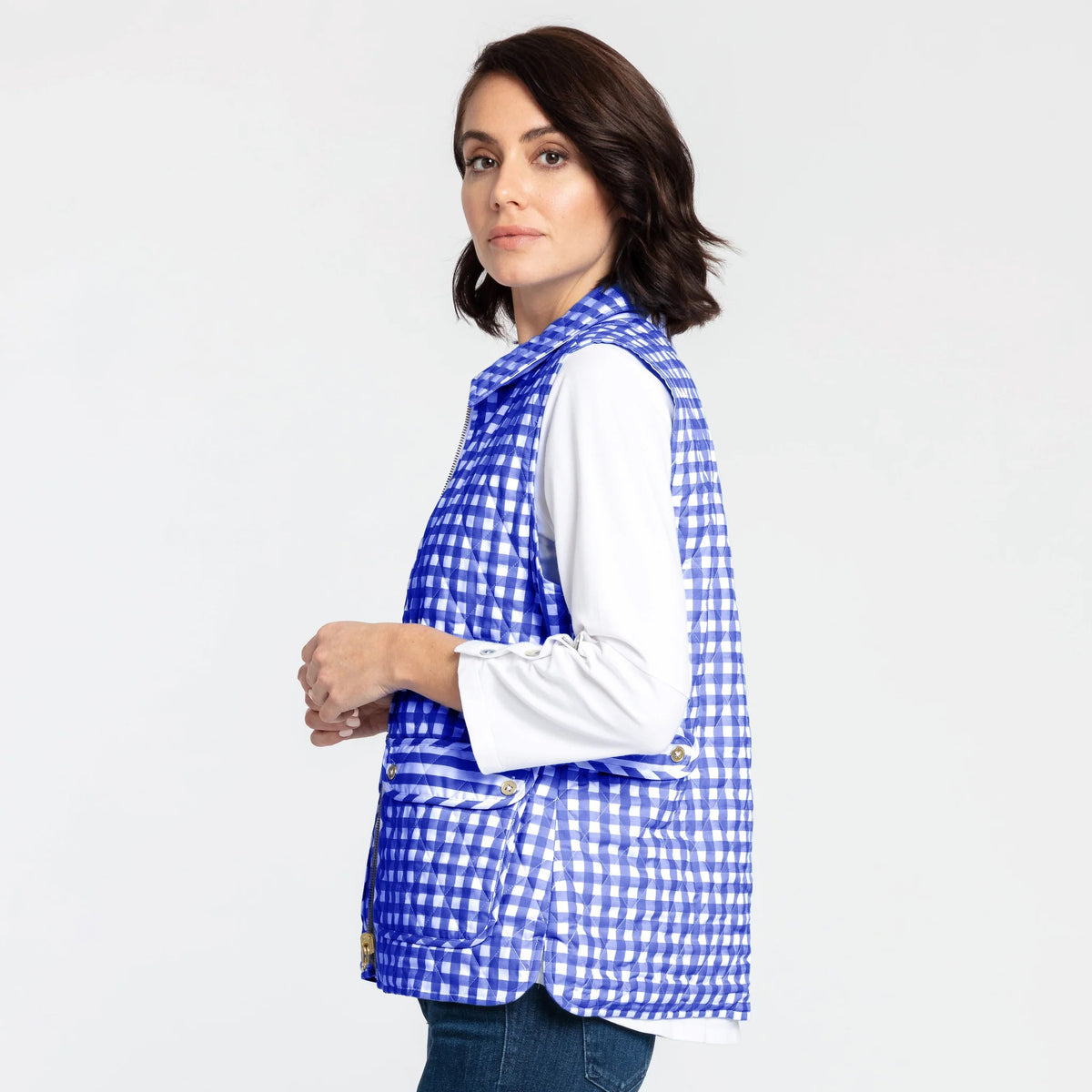 A woman with shoulder-length dark hair is wearing the HINSON WU - LAUREN VEST, which is fully reversible and features a blue and white gingham pattern, over a white long-sleeve shirt. She is standing sideways, looking at the camera with her left hand resting near her waist. The background is plain white.