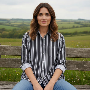 A woman with wavy brown hair sits on a wooden bench outdoors, surrounded by green fields and hills beneath a cloudy sky. She is wearing blue jeans and the FRANK & EILEEN EILEEN Relaxed Button Up in Italian Brushed Superluxe Cotton Black and Tan Stripe by FRANK & EILEEN.