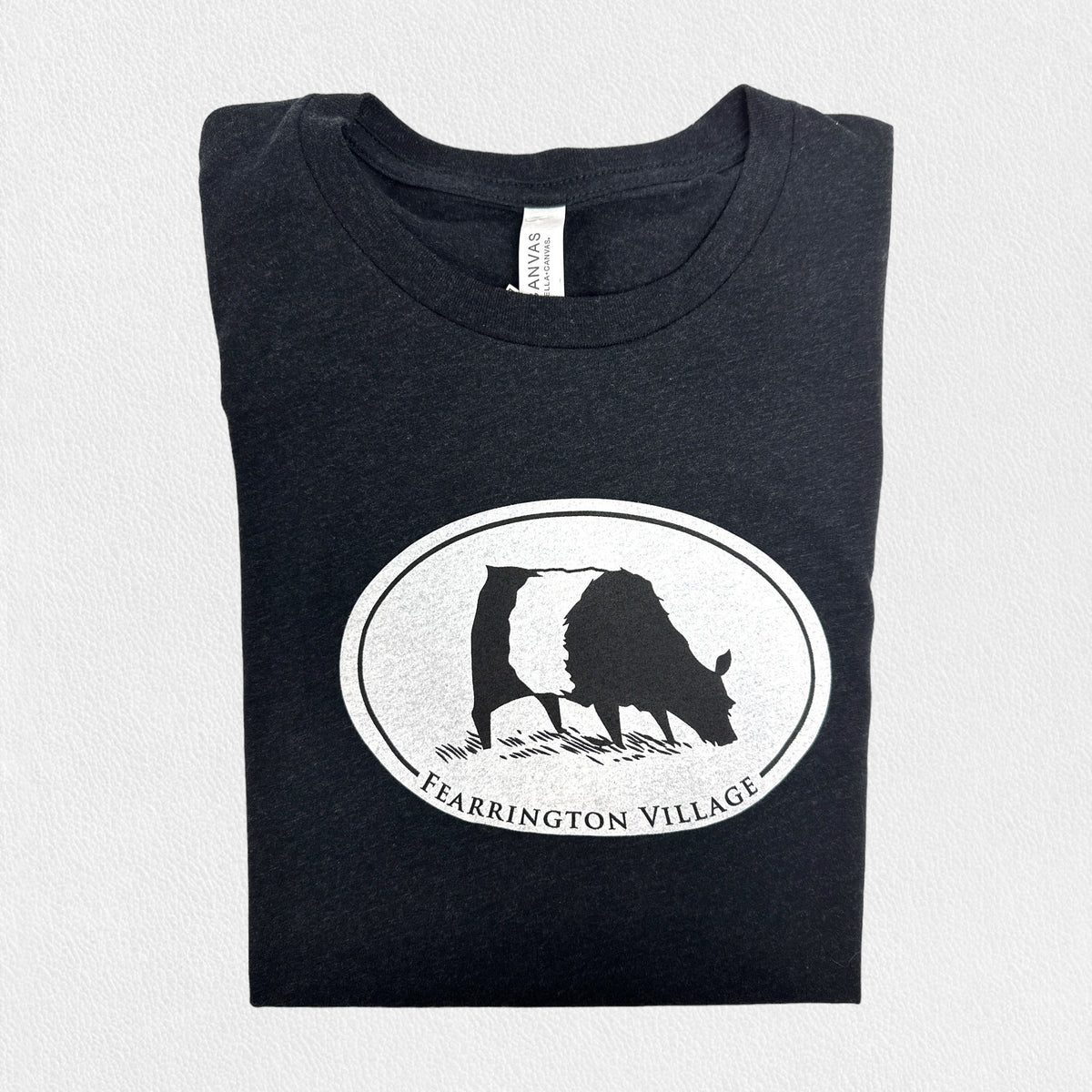 A black triblend unisex tee from the FEARRINGTON LIFESTYLE COLLECTION by GILDAN, showcasing a white oval logo with a silhouette of a goat and the text &quot;FEARRINGTON VILLAGE&quot; underneath, set against a plain light gray background.