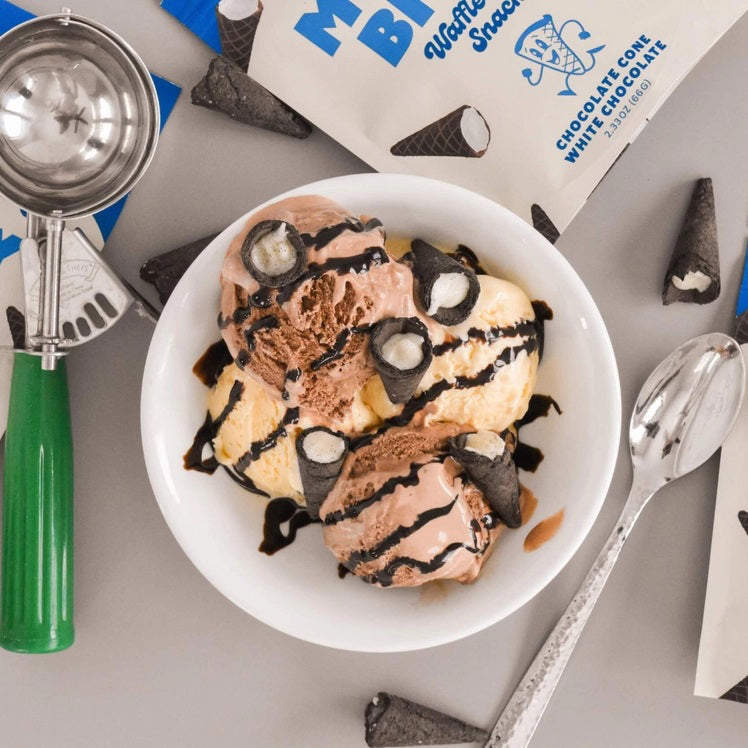 A person dressed in a light blue outfit sits on a plush, royal blue rug holding a bag of Muddy Bites Cookies &amp; Cream Waffle Cones in one hand and a small white chocolate waffle cone snack in the other. The beige and blue bag features an illustration of chocolate-dipped waffle cones.