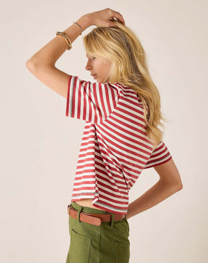 A smiling person with long blonde hair wears the MERSEA Deauville Ringer Tee in Currant Jade—a red and white striped ringer tee with green collar and cuffs—paired with green pants, standing against a light background.