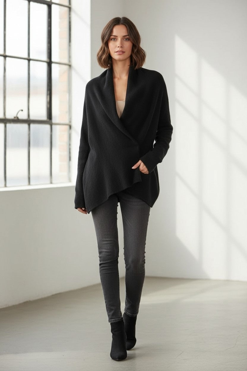 A woman stands indoors by large windows, wearing the JESSIE LIU - KNIT JACKET WITH ASYMMETRICAL CLOSURE, styled with gray skinny jeans and black ankle boots. Natural light fills the minimalist room with white walls and a concrete floor.