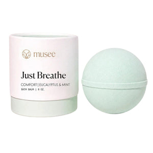 A pale green sphere infused with essential oils rests beside a white cylindrical container labeled “MUSEE - JUST BREATHE THERAPY BATH BALM,” inviting you to unwind into a spa-like escape.