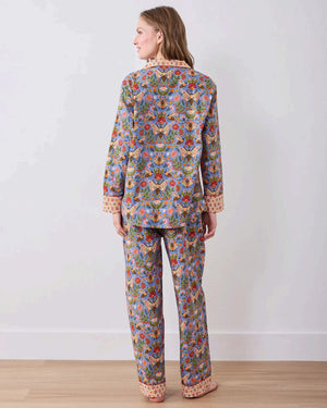A woman with light brown hair sits against a white wall, wearing the PRINTFRESH - Bee Tapestry Long Pajama Set in Delphinium Blue. This organic cotton pajama set from PRINTFRESH features a colorful floral and bee pattern with vibrant red and yellow cuffs, collar, and pocket trim, beautifully blending playful design with cozy comfort.