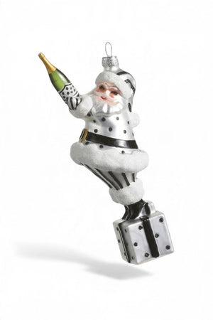 A CHAMPAGNE POPPING SANTA ORNAMENT by RAZ Imports is a resin Santa Claus wearing a silver and black outfit, holding a champagne bottle in one hand and standing on a gift box.