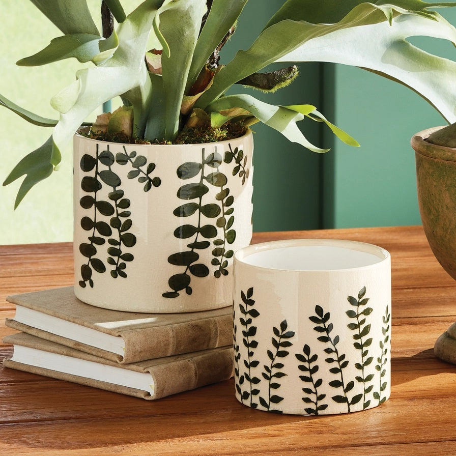 Two NAPA HOME AND GARDEN Maren Pots in white ceramic with an embossed leaf pattern sit on a wooden table, one with a leafy plant. Nearby are two stacked books and a partially visible empty green cachepot.