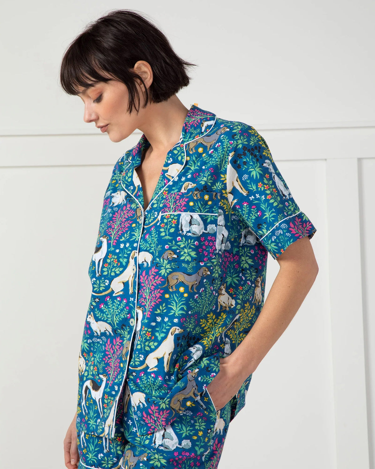 A person stands against a plain white wall wearing the stylish PRINTFRESH - MUST LOVE DOGS PAJAMA SHORT SET made of organic cotton. The set, adorned with colorful dog and floral illustrations on a blue background, features pockets where the person&#39;s hands rest.