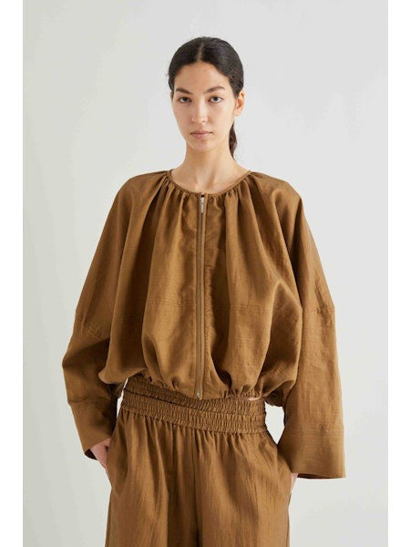 A person with long, dark hair in a low ponytail, held by a silver clip, is seen from behind wearing the MEIMEIJ - COTTON BOMBER JACKET in brown. The background is plain white.