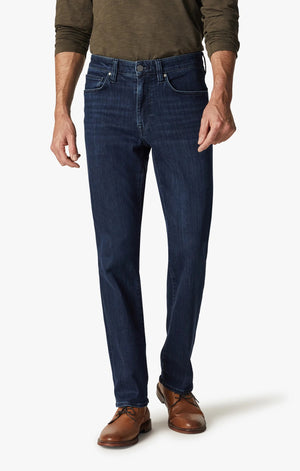 A man in a green long-sleeve shirt, brown shoes, and 34 HERITAGE Charisma Relaxed Straight Leg Jean in Dark Midnight Brushed Urban wash stands with his back to the camera against a plain white background.