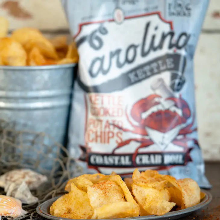 A light blue bag with a rustic wooden plank design labeled &quot;CAROLINA KETTLE - CRAB BOIL CHIPS 5OZ&quot; from the brand &quot;1 IN 6 SNACKS&quot;. Featuring a red crab holding a map, the text &quot;Certified Awesome&quot; and &quot;Guaranteed Fressher,&quot; 5 oz (141.8g), GF, and the &quot;1 in 6 Snacks&quot; logos. Enjoy this savory treat inspired by Southern tradition.