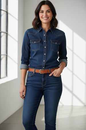 A woman stands indoors by a window, smiling and wearing the MERSEA MARGOT BUTTON UP SHIRT from MERSEA, featuring pearl snaps and a tailored fit with rolled-up sleeves. She pairs the dark denim shirt with matching jeans and a brown belt, while her dark hair falls in loose waves.