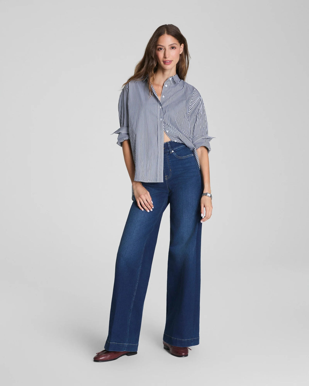 A woman with long brown hair stands against a plain background, smiling slightly. She is wearing a blue and white striped blouse paired with the SPANX Shape EveryWear Pull On Wide Leg Jean in Poseidon by SPANX and brown shoes, accessorized with a watch on her wrist.