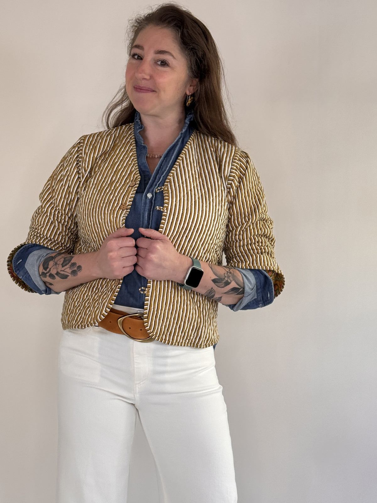 A woman with visible forearm tattoos smiles slightly against a plain background, wearing the FARYLINA ZOIE Reversible Quilted Jacket over a denim shirt, white pants, and a tan belt.
