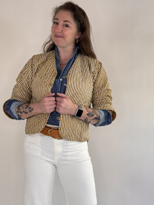 A woman with visible forearm tattoos smiles slightly against a plain background, wearing the FARYLINA ZOIE Reversible Quilted Jacket over a denim shirt, white pants, and a tan belt.