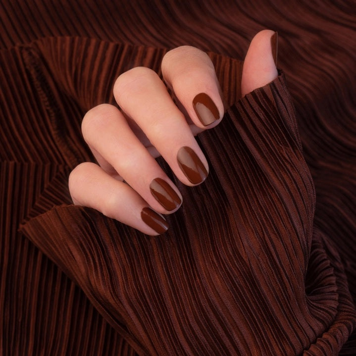 A hand adorned with MANUCURIST - GREEN™ Nail Polish in Chestnut by MANUCURIST INC rests on a wooden table beside a cup of coffee with heart-shaped latte art and a small pastry sprinkled with coarse sugar.