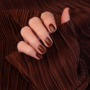 A hand adorned with MANUCURIST - GREEN™ Nail Polish in Chestnut by MANUCURIST INC rests on a wooden table beside a cup of coffee with heart-shaped latte art and a small pastry sprinkled with coarse sugar.