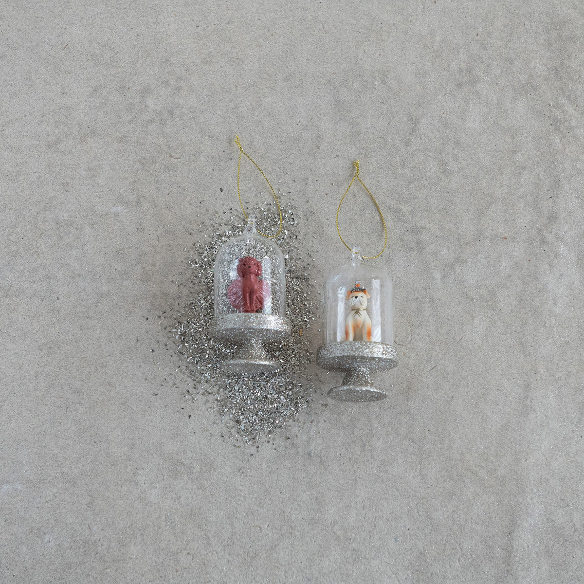 The CREATIVE COOP DOG IN CLOCHE ORNAMENT set includes two glass ornaments with gold bases and loops: one features a crowned dog, the other a pink poodle, each displayed on separate stands against a white background.