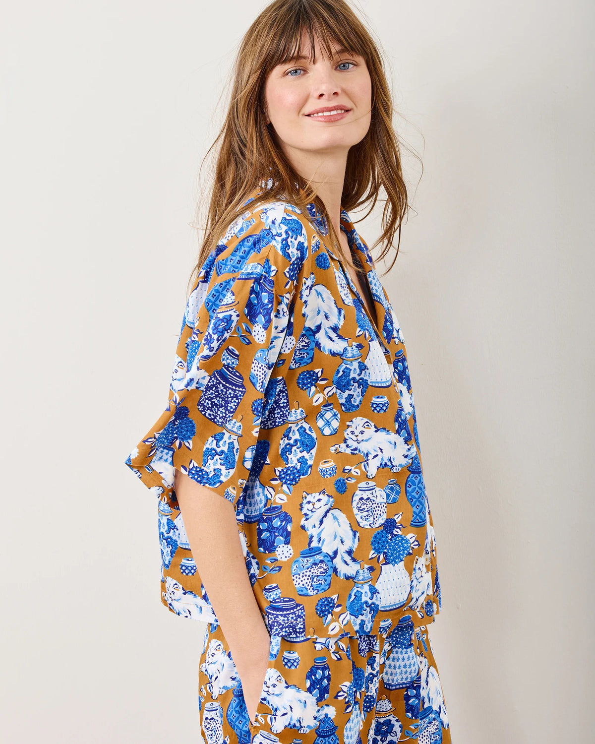 A person in Printfresh&#39;s Satin Fancy Cat Wildest Dreams pajama set, crafted from eco-friendly viscose and adorned with blue, white, and brown hand screen-printed motifs, stands barefoot by a plain wall. They&#39;re smiling with one hand touching their chin.