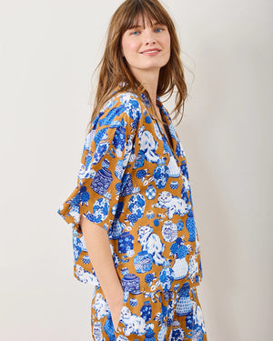 A person in Printfresh's Satin Fancy Cat Wildest Dreams pajama set, crafted from eco-friendly viscose and adorned with blue, white, and brown hand screen-printed motifs, stands barefoot by a plain wall. They're smiling with one hand touching their chin.