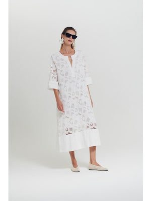 A woman in the MILVA MI Embroidered Cotton Dress in Black by MILVA, featuring three-quarter sleeves and subtle embroidery, stands against a white background. She has short hair, sunglasses, white earrings, and flat shoes.