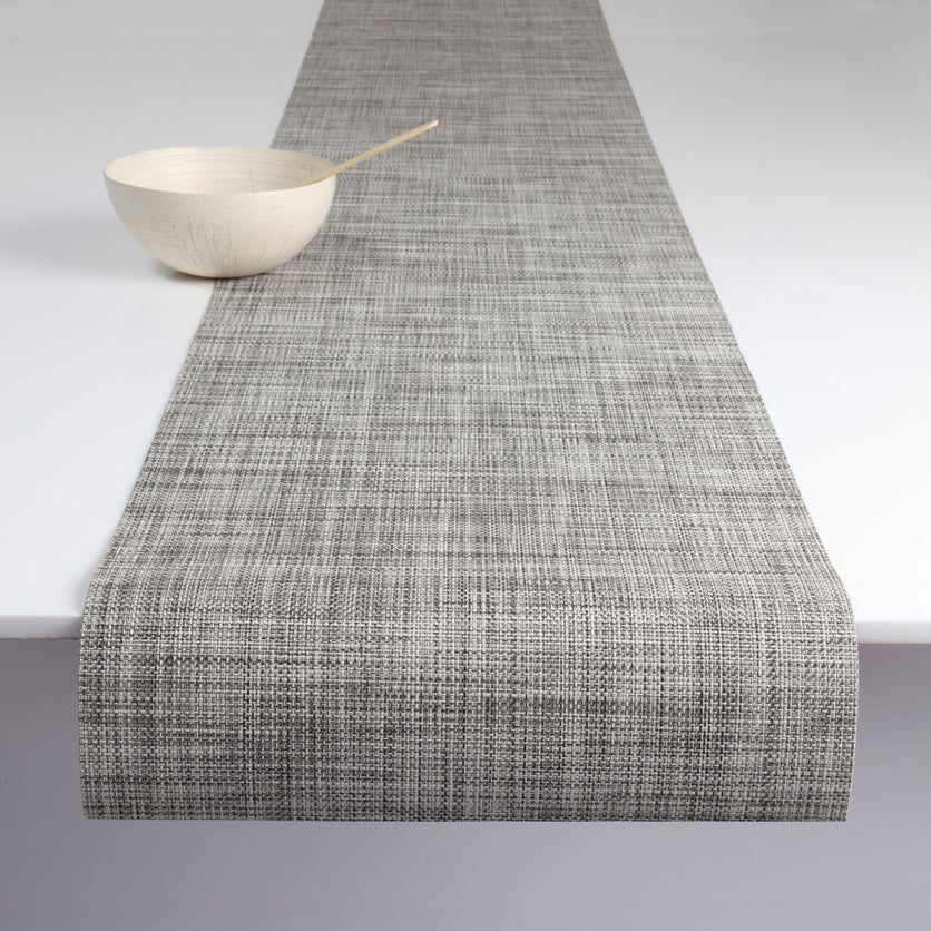 The CHILEWICH MINI BASKETWEAVE RUNNER 2.5X8 in textured gray covers a white table, accented by a light wood bowl and matching spoon at one end.